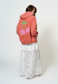 Koraalkleurige hoodie met grafische tekst in pink en groen op de achterkant, gecombineerd met een witte maxi-jurk van kant met een bloemenpatroon.