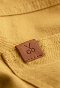 Étiquette en cuir marron avec une illustration de ciseaux cousue sur un tissu texturé moutarde, présentant des coutures visibles et un détail de surface lisse.