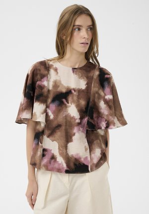 Žena so svetlohnedými vlasmi, oblečená v voľnom top s krátkym rukávom s hnedými, čiernymi, krémovými a ružovými abstraktnými vzormi a krémovými nohavicami.