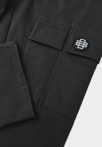 Zwarte cargobroek met meerdere zakken en een gestructureerde stof. Opvallend detail is een contrasterend logo-patch in marineblauw en zilver.