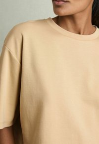 Yksinkertainen beige oversize T-paita, jossa on pyöreä kaula-aukko, lyhyet hihat ja pehmeä, sileä rakenne. Ei näkyviä kuvioita tai metallisia yksityiskohtia.