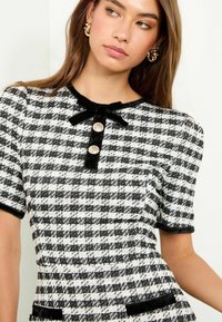 Mustvalge houndstooth mustriga kleit lühikeste varrukatega, sametist ääristusega, paelaga detailiga ja kahe dekoratiivnupuga kaeluse juures.