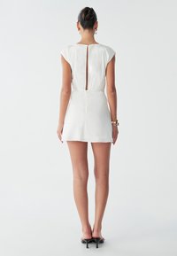 Hvid kjole med nøglehuldetalje bagpå, kapærmer og en figursyet talje. Kort nederdel med en glat tekstur. Kombineret med sorte hælede sandaler.