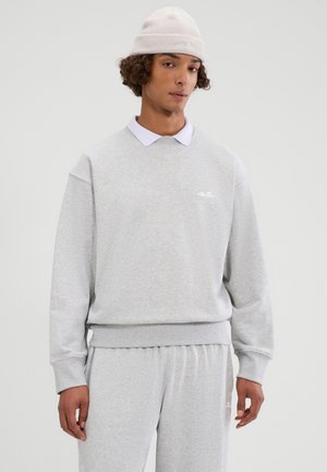 Jonge man met een lichtgrijze sweatvest en bijpassende joggingbroek, een wit overhemd met kraag eronder, en een lichtroze gebreide muts.