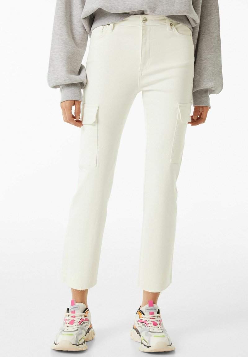 Pantalon cargo blanc à taille haute, avec deux poches latérales, tissu texturé et ourlets effilochés, associé à des baskets multicolores.