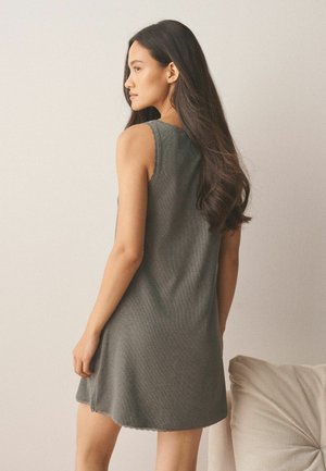 Woman with long dark hair wearing a sleeveless gray dress stands facing sideways near a beige pillow on a light background.