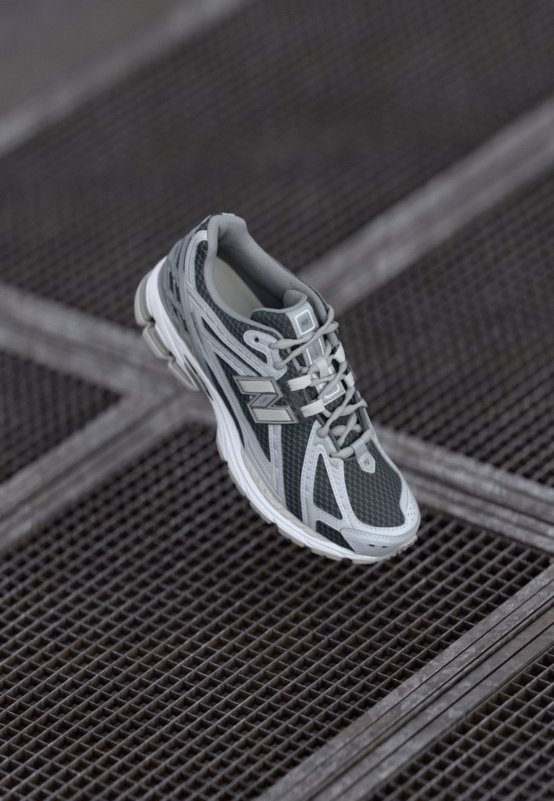 Chaussure de running New Balance grise et blanche suspendue au-dessus d'une surface en grille métallique avec des lignes croisées en arrière-plan.