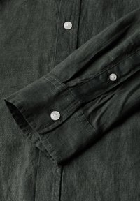 Manica di camicia in lino verde scuro con polsino a bottoni, con una sottile texture e dettagli di cucitura visibili. I bottoni sono rotondi e bianchi.