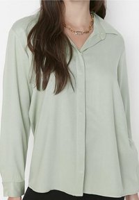 Light green, long-sleeve button-up shirt with a soft texture, collared design, and subtle sheen. Gold chain necklace visible.