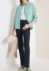 Femme portant une chemise à boutons vert menthe sur un t-shirt blanc, jean bleu foncé, tenant une veste beige, et portant des baskets blanches et vertes.