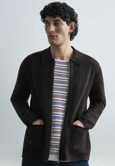 Veste marron à fermeture éclair avec tissu texturé, deux poches avant et un col à revers, portée sur un pull à motifs avec des rayures bleues, rouges et crème.