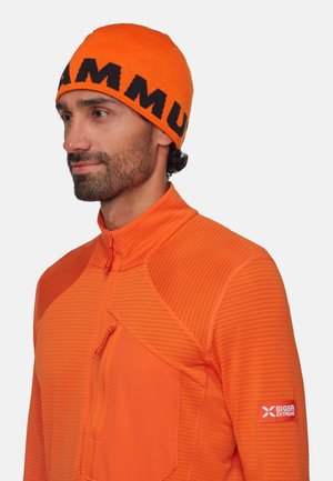 Orange knit beanie with black "MAMMUT" lettering, worn with an orange zip-up jacket featuring textured and smooth fabric sections.