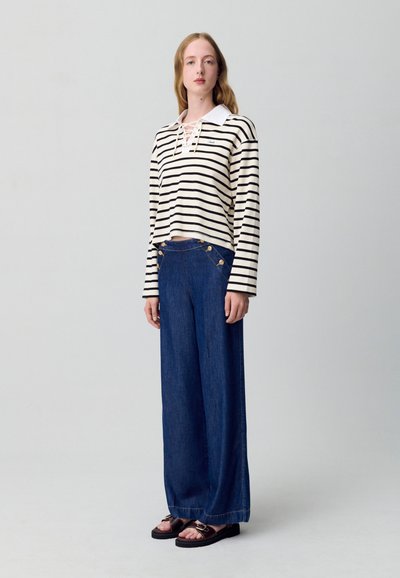 Mujer vistiendo una blusa de rayas blanca y negra de manga larga con collar con cordones, pantalones vaqueros de pernera ancha azul con botones dorados y sandalias negras.