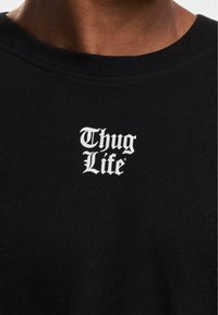 Fekete bordázott pamut póló, az elején "Thug Life" felirattal fehér gótikus stílusú betűkkel, kerek nyakkal és testhez simuló fazonnal.