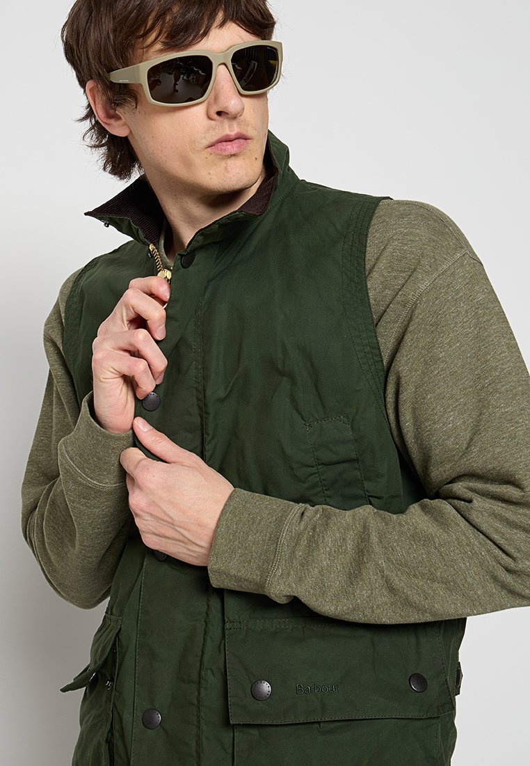 Mann mit Sonnenbrille und einer grünen gepolsterten Weste über einem olivfarbenen Langarmshirt, der den Reißverschlusskragen der Weste anpasst.