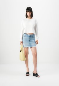 Kvinne som står, iført hvit langermet crop topp, slitt denim miniskjørt, svarte kilehæler, holder en stor lys gul håndveske.