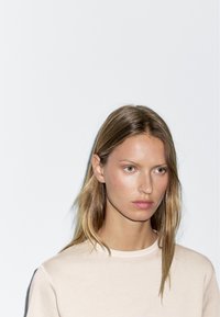 Ein schlichter, beiger Langarmshirt mit rundem Ausschnitt, getragen von einer Person mit schulterlangen braunen Haaren, vor einem weißen Hintergrund.