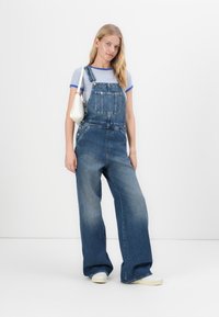 Jeans-Latzhose in mittlerer Waschung, mit weitem Bein, ausgestattet mit Fronttaschen und Knopfverschlüssen. Kombiniert mit einem blau gestreiften T-Shirt und einer weißen Tasche.