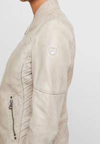 Světlé béžové kožené sako s vroubkovaným bočním designem, přední kapsou na zip a malou nášivkou loga na rameni. Hladký povrch.