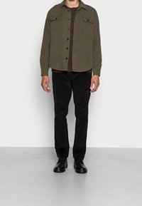 Camisa abotonada verde oliva con dos bolsillos en el pecho, usada sobre un suéter marrón acanalado, combinada con pantalones negros y zapatos negros.