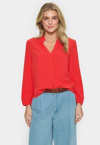 Blusa rossa a maniche lunghe con scollatura a V e colletto arricciato, abbinata a pantaloni blu a vita alta e una cintura marrone. Tessuto morbido, vestibilità casual.