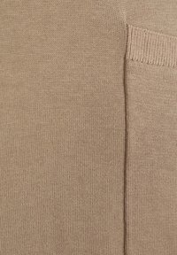 Gebreide trui in tan kleur, met een geribbelde halslijn en een zakdetail. De textuur is zacht en consistent over de stof.