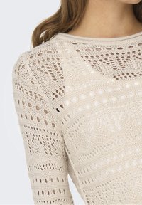 Pull-over beige en maille présentant des motifs ajourés complexes, un col rond et des manches longues avec des détails texturés.
