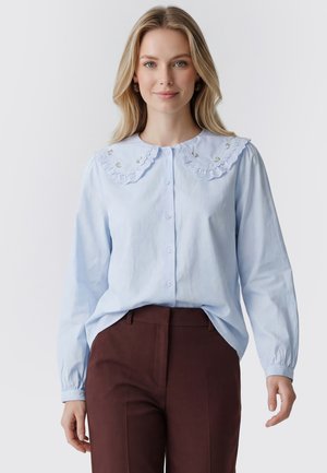 Kvinde med langt blond hår iført lyseblå bluse med broderet, flæsekrave og brune bukser, stående mod en ensfarvet baggrund.