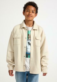 Giacca oversized color beige chiaro con due tasche sul petto, abbinata a una t-shirt grafica con design di un gatto e jeans blu. Tessuto morbido.