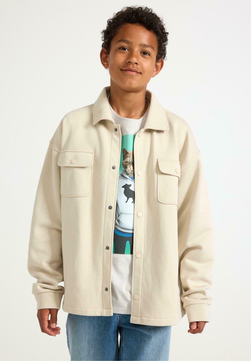Giacca oversized color beige chiaro con due tasche sul petto, abbinata a una t-shirt grafica con design di un gatto e jeans blu. Tessuto morbido.
