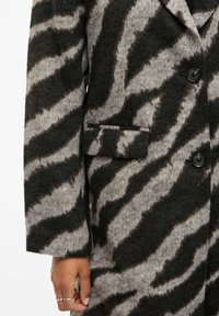 Nærbillede af en hånd, der bærer sølvringe, ved siden af en brun og grå zebra-stribet uldfrakke med knapper og en klaplomme.