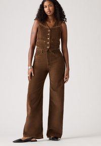 Bruine corduroy gilet met knopen aan de voorkant, gecombineerd met hooggeknoopte bruine corduroy wijde broek. Model draagt zwarte puntige flats.