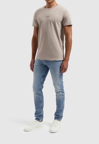 Beige katoenen t-shirt met ronde hals en klein zwart logo op de borst, gecombineerd met lichtblauwe spijkerbroek en witte sneakers.
