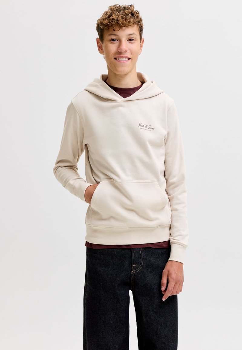 Jeune homme aux cheveux bouclés portant un sweat à capuche crème avec poche avant et un jean foncé, debout devant un fond clair uni.