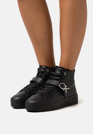 Baskets montantes noires à semelles épaisses, lacets à l'avant, bride avec logo argenté "CK", portées sur jambes nues sur fond blanc.