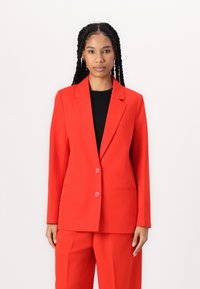 Rode blazer en bijpassende broek gemaakt van een gladde stof. De blazer heeft een omgeslagen kraag, twee knopen en voorzakken.