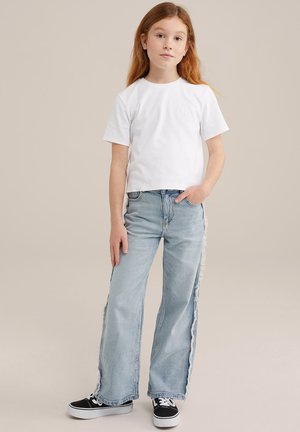 camiseta blanca de manga corta con un pequeño detalle bordado, combinada con unos vaqueros de pierna ancha azul claro con costuras laterales deshilachadas. Zapatillas negras.