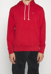Homme portant un sweat à capuche rouge Levi's avec des cordons blancs et un pantalon en velours côtelé foncé, debout devant un fond blanc uni.