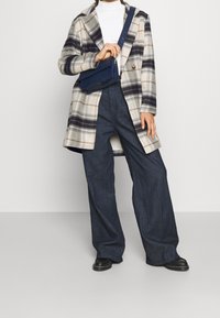 Rutig ullblandad kappa i beige och marinblå, tillsammans med mörkblå vida byxor. Accenterad med en marinblå väskan i faux-krokodil som bärs över axeln.