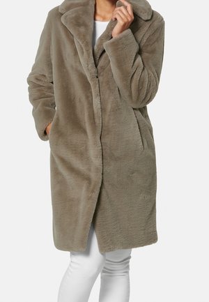 Vrouw draagt een taupekleurige faux fur jas van midilengte met een brede kraag, zakken en lange mouwen, gecombineerd met een witte broek.