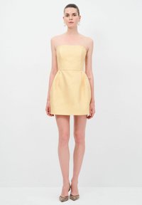 Robe jaune sans manches avec un corsage ajusté, une jupe évasée, un tissu texturé et des poches latérales ; associée à des talons à bouts pointus ornés.