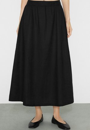 Kobieta ubrana w czarną spódnicę do kostek z elastyczną gumką w pasie i czarne płaskie buty, stojąca na szarej podłodze.