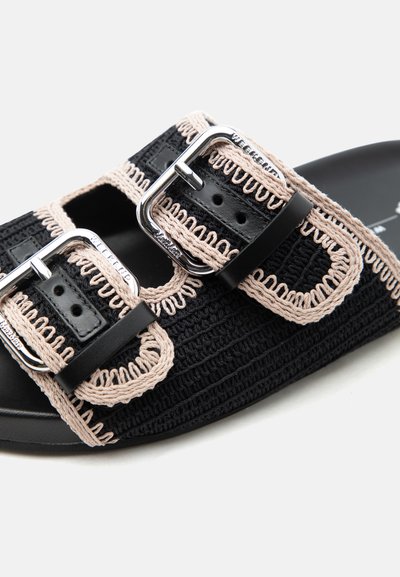 Μαύρο ανοιχτό σανδάλι με δύο μπεζ ραμμένες υφασμάτινες λουρίδες που διαθέτουν ασημί ρυθμιζόμενες μεταλλικές αγκράφες, σε ανοιχτό φόντο.