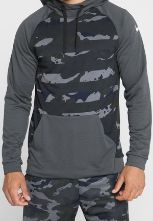 Mann trägt dunkelgrauen und schwarzen Hoodie mit Camouflage-Muster und passende Camouflage-Hose, steht mit locker an den Seiten hängenden Händen.