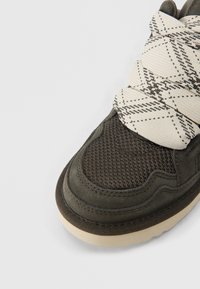 Σκούρο ελιάς πράσινο αθλητικό παπούτσι με υφασμάτινη υφή στη μύτη, σουέντ λεπτομέρειες, χοντρές κρεμ κορδέλες με μαύρα γεωμετρικά σχέδια και λευκή σόλα.