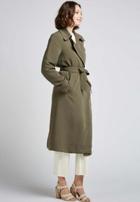 Long manteau vert olive avec une taille ceinturée, un large col et des poches latérales, associé à un pantalon blanc cropped et des sandales compensées beiges.
