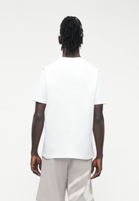 Juodaodis asmuo su dredais, dėvintis paprastą baltą trumpomis rankovėmis marškinėlį ir šviesiai pilkas šortus su balta detale, stovintis nugara.