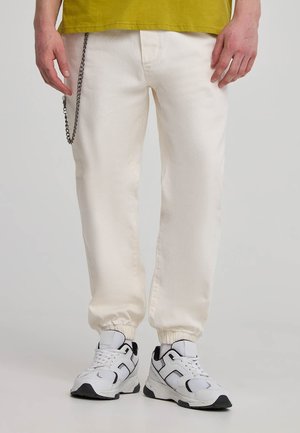 Pantaloni jogger bianchi in cotone, con polsini elasticizzati, un dettaglio in catena laterale e abbinati a sneakers nere e bianche.