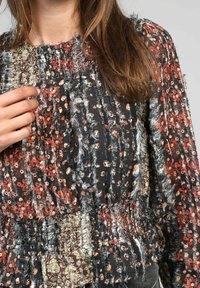 Gros plan sur une personne portant un haut floral texturé multicolore avec des nuances de noir, rouge, orange et beige, la main touchant la zone de la poitrine.