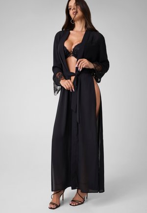 Femme portant une longue robe noire transparente avec manches en dentelle sur un soutien-gorge noir, nouée à la taille, debout en talons noirs à brides sur fond gris.